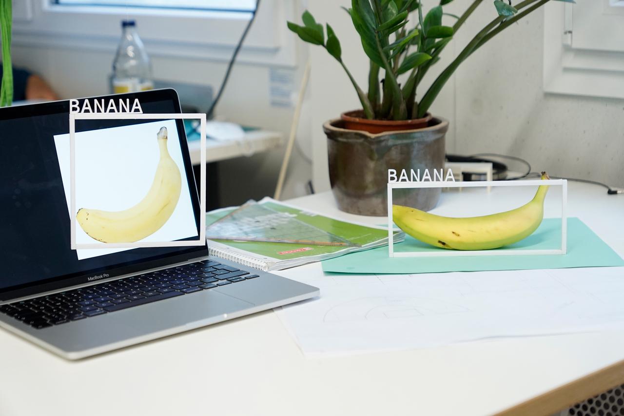 A banana on a table and an image of a banana on a laptop on the same table. Each of the two bananas has a white frame around it with the word 'Banana' sticked on top of it