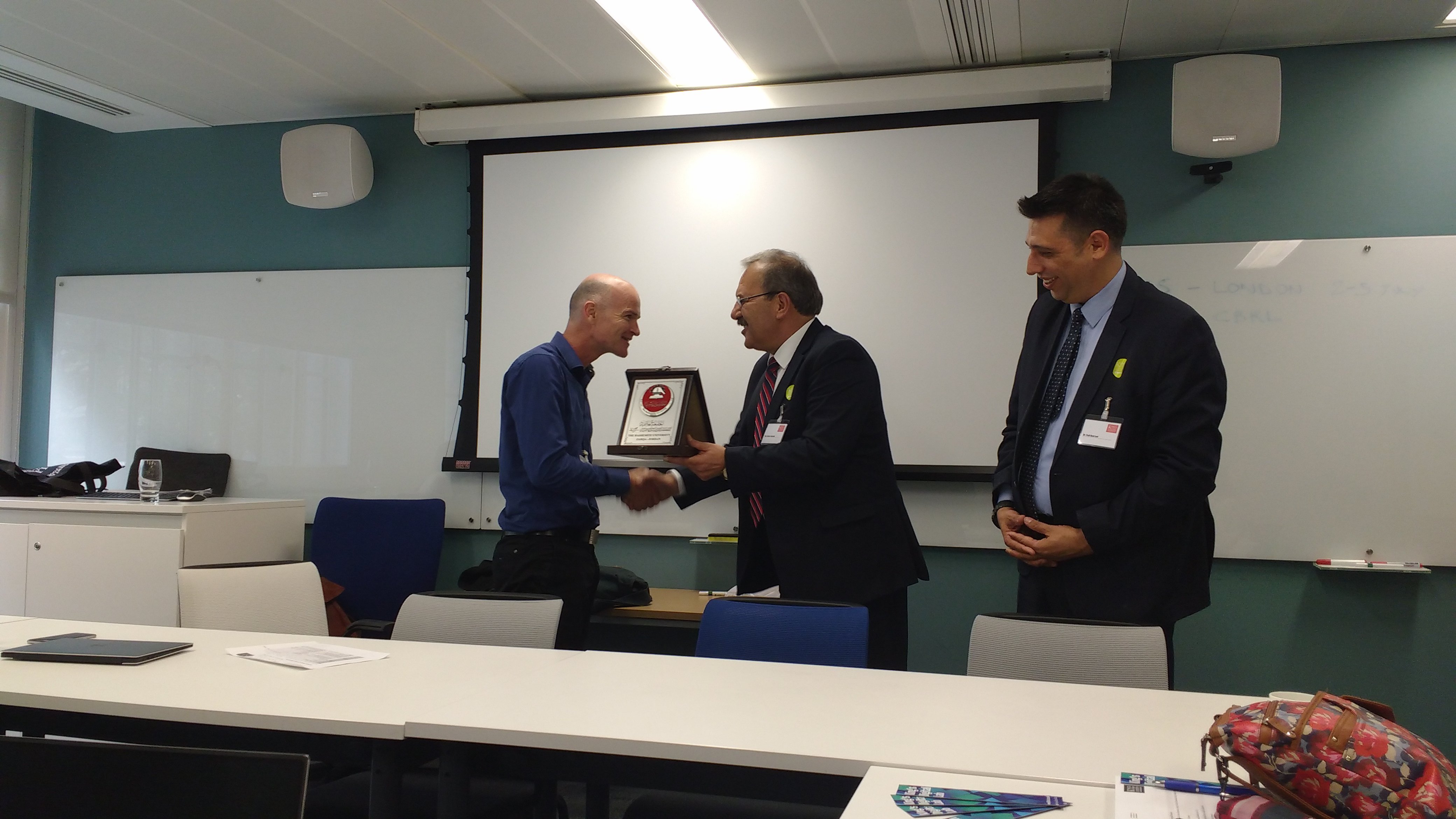 Three men in a meeting room, with one man receiving an award from another man while the man on the left watches with a smile