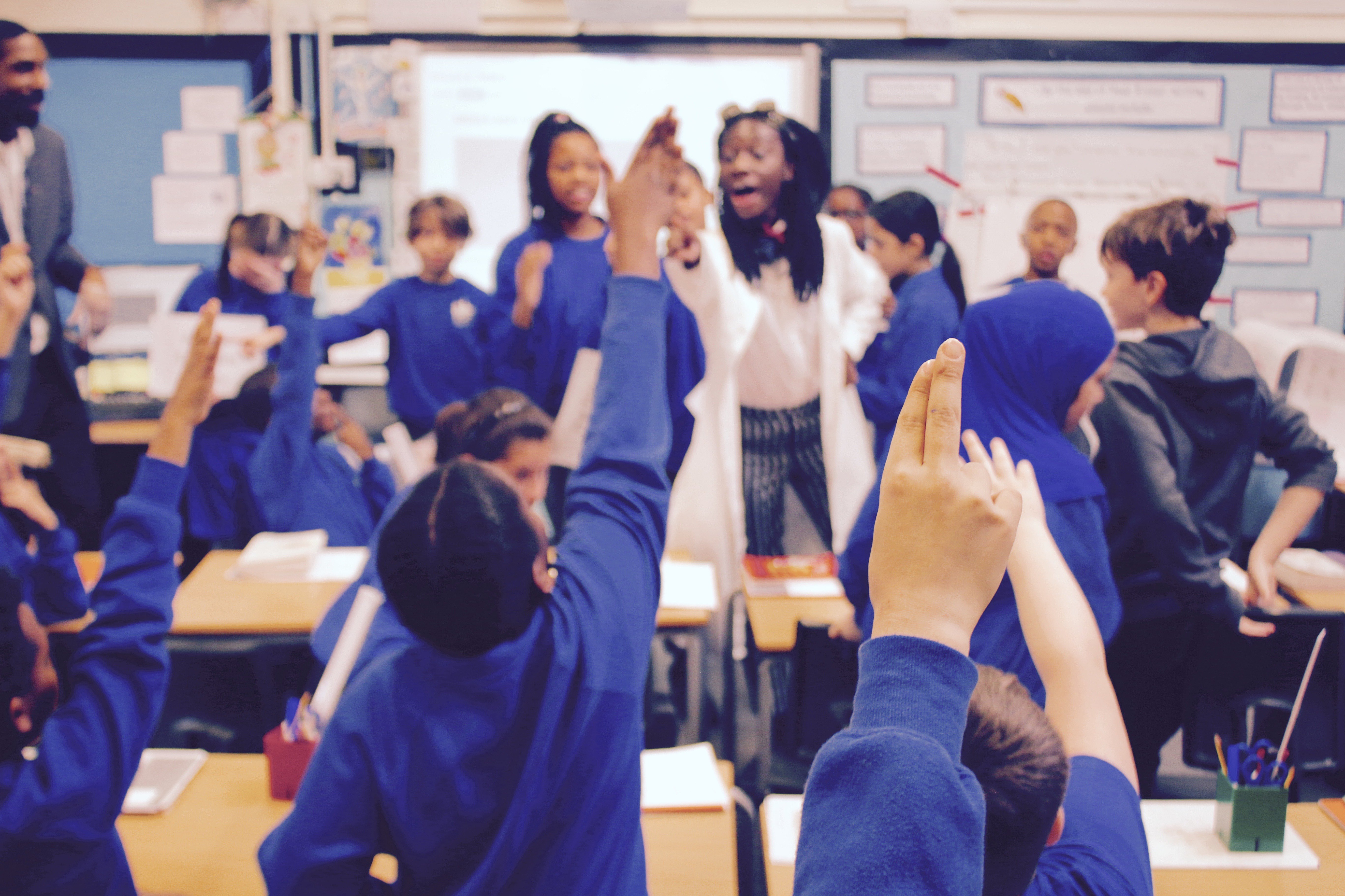 A lively classroom scene with students in blue uniforms standing and raising their hands