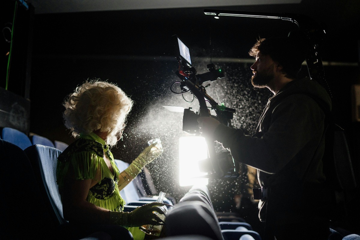 A filmmaker records a performer in a bright green costume under dramatic lighting inside a dark theatre.