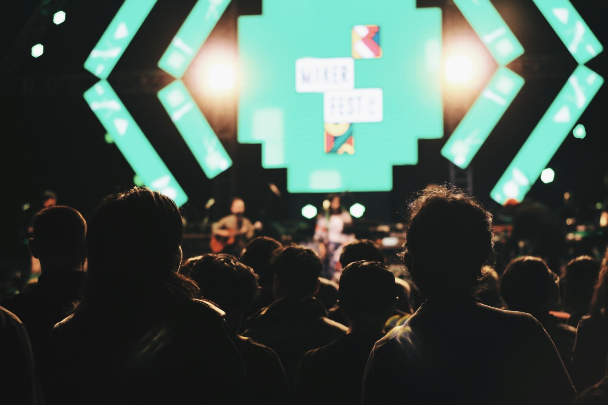 A crowd watching a live performance on a brightly lit stage with a large digital screen in the background.