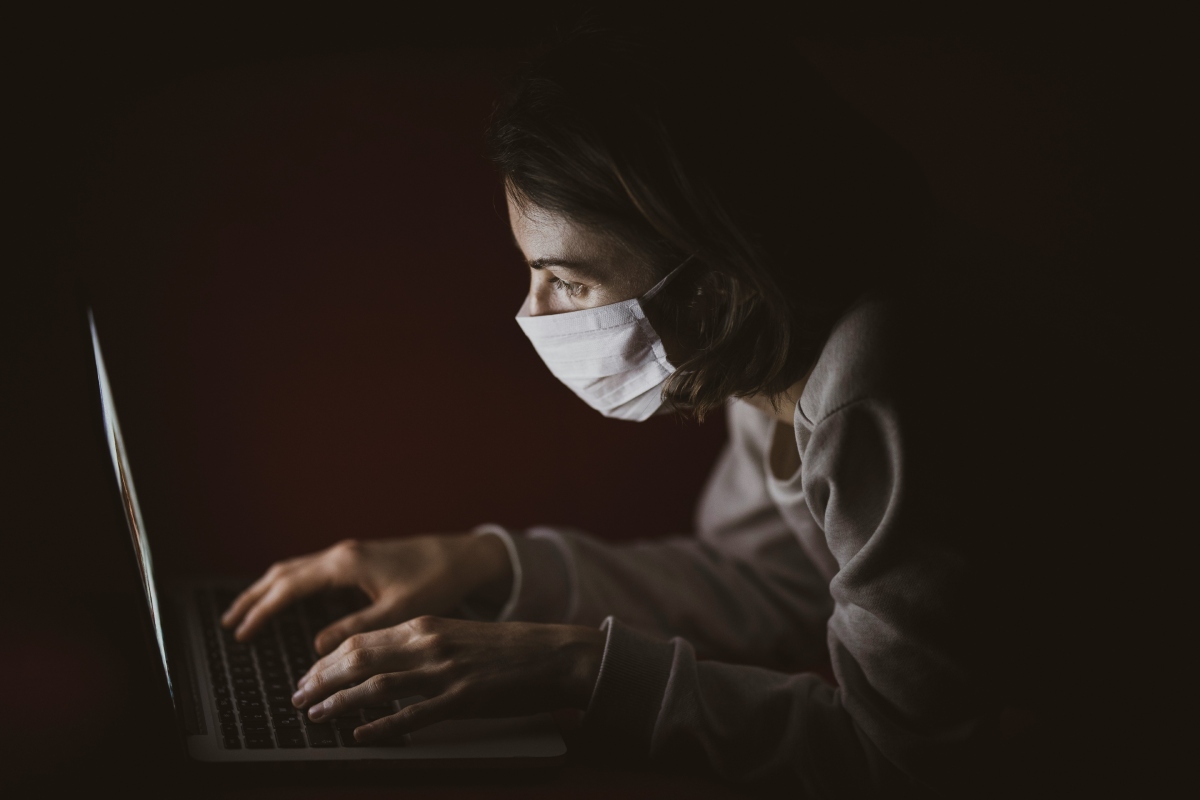 A person wearing a face mask, hunched over a laptop in a dimly lit room, their face illuminated by the screen’s glow.