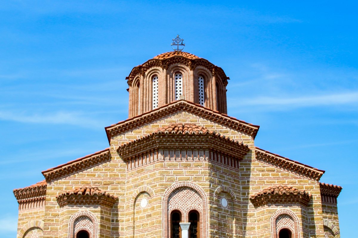 A Byzantine-style church with intricate brickwork, arched windows, and a central dome topped with a cross, set against a clear blue sky.