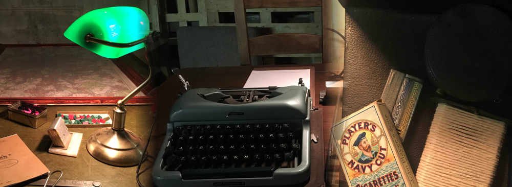 Vintage workspace corner featuring an antique typewriter, a green lamp, a cigarette box and small personal artifacts.