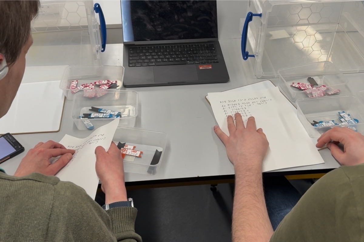 Two people sit at a table sorting printed code strips into clear containers, with a laptop open in front of them.