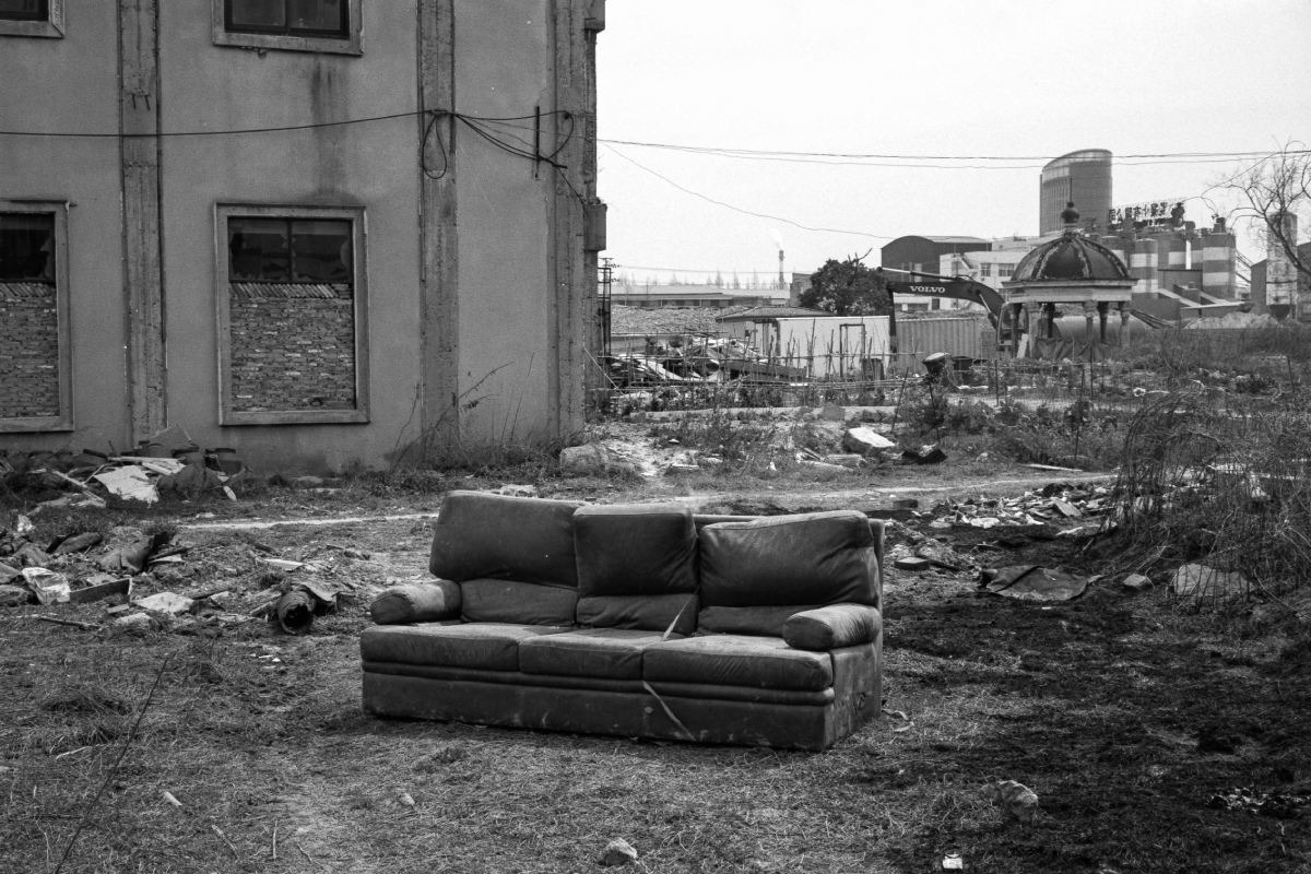 An abandoned sofa sits in an empty, overgrown lot beside a damaged building, with construction machinery and industrial structures in the background.