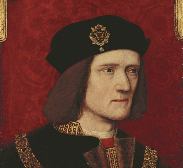 A head and shoulders portrait of Richard III, on a red background with gold embellishment in the upper corners. His face is turned slightly to the right, with a serious expression; at the bottom of the frame, his hands are shown in the middle of removing a ring from his right little finger. The heights of his shoulders are visibly uneven. He wears a black robe, possibly fur-lined, over a gold patterned doublet with a flash of red at the collar. A chain of office hangs across his chest. His black hat bears a gold, ruby, and pearl brooch in the shape of a stylised rose.
