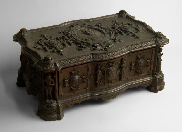 A wooden box with a hinged lid, varnished to a dark mahogany colour. The surfaces have decorative carvings. In each corner, a figure representing a character from Shakespeare's plays stands in an alcove. On the side of the box facing the viewer, a coat of arms is carved on the centre panel, with medallions either side embossed with Shakespeare's entwined initials. On the lid, a portrait of Shakespeare in a decorative oval in the centre is flanked by two smaller shields carved with his birth date of 23 April 1564 and death date of 23 April 1616.