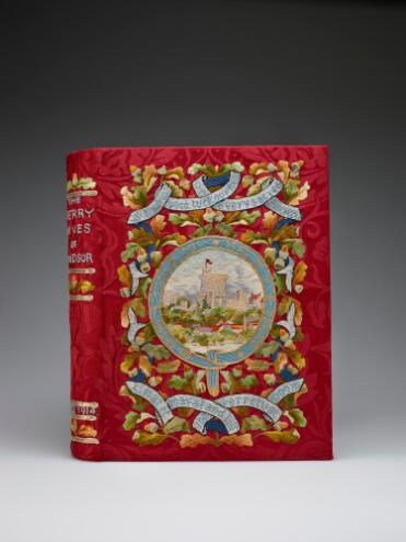 A book with a colourful embroidered cover. The background is a red brocade silk. At the centre of the cover a round view of Windsor Castle is framed by the blue ribbon of the Order of the Garter, embroidered with its motto 'Honi soit qui mal y sense.' Above and below are further ribbons bearing quotes from the play ('Strew good luck ouphes in every sacred room/ That it may stand till the perpetual doom'), strung across a backdrop of oak leaves and acorns. On the book's spine, the title 'The Merry Wives of Windsor' can be seen embroidered in blue thread, and the date A.D. 1914, with further oak leaf and acorn motifs.