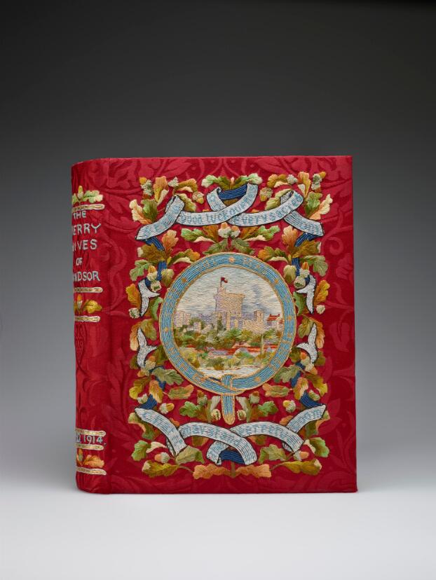 A book with a colourful embroidered cover. The background is a red brocade silk. At the centre of the cover a round view of Windsor Castle is framed by the blue ribbon of the Order of the Garter, embroidered with its motto 'Honi soit qui mal y sense.' Above and below are further ribbons bearing quotes from the play ('Strew good luck ouphes in every sacred room/ That it may stand till the perpetual doom'), strung across a backdrop of oak leaves and acorns. On the book's spine, the title 'The Merry Wives of Windsor' can be seen embroidered in blue thread, and the date A.D. 1914, with further oak leaf and acorn motifs.